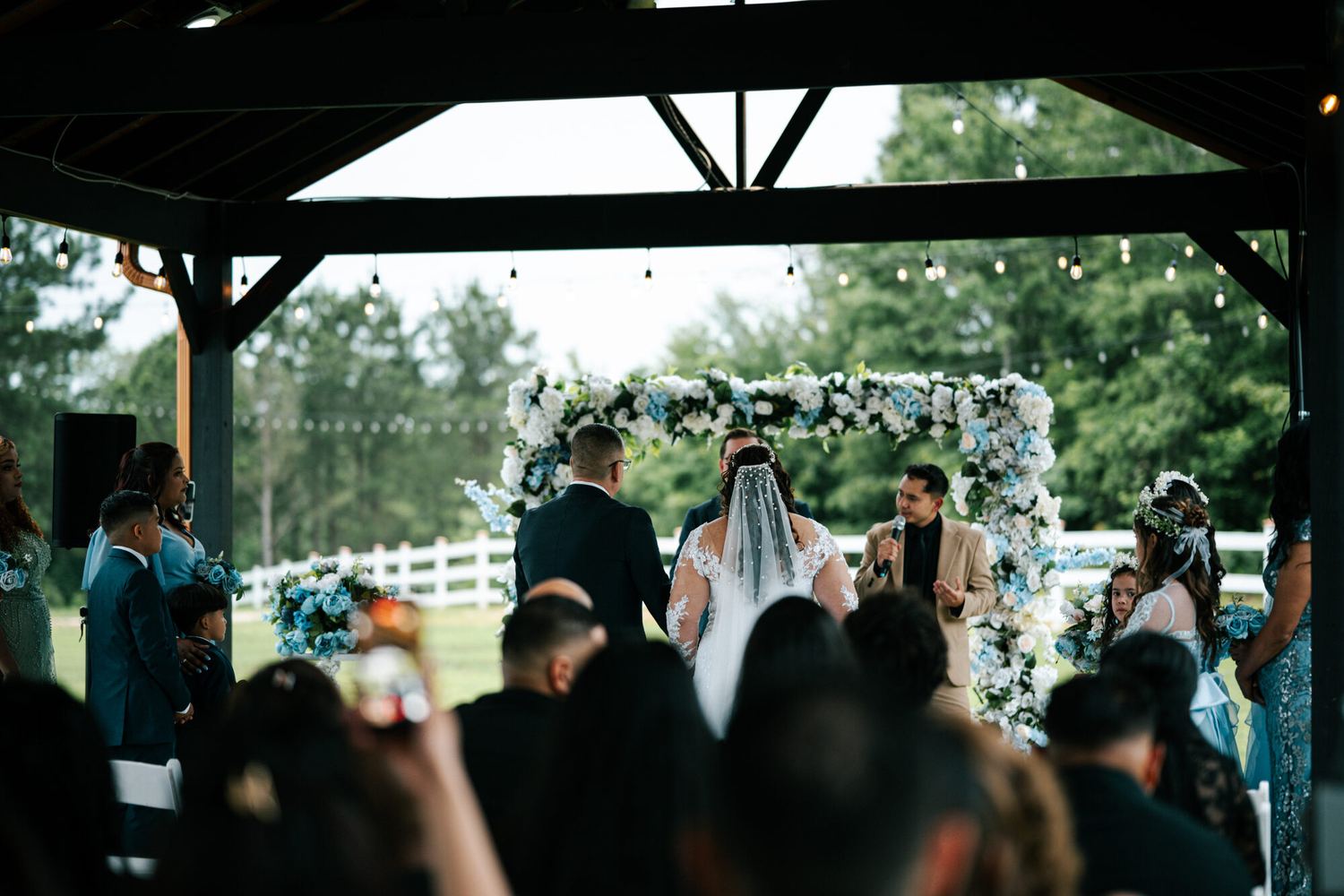 Ceremonia de boda al aire libre bajo un arco de flores azules y blancas, con luces colgantes